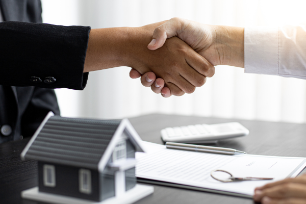 wei Personen schütteln sich die Hand über einem Vertrag. Im Vordergrund steht ein kleines Modell eines Hauses, daneben liegen ein Stift und eine Brille.