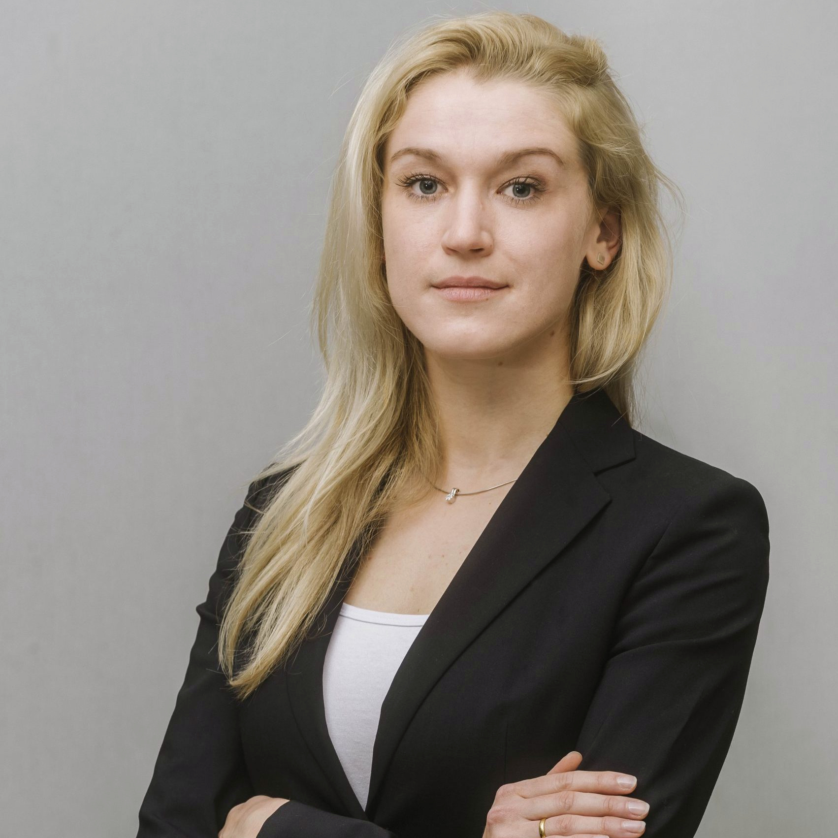 Mag. Julia Hertzberg, eine Frau mit blondem Haar, trägt einen schwarzen Blazer und posiert vor einem neutralen Hintergrund.